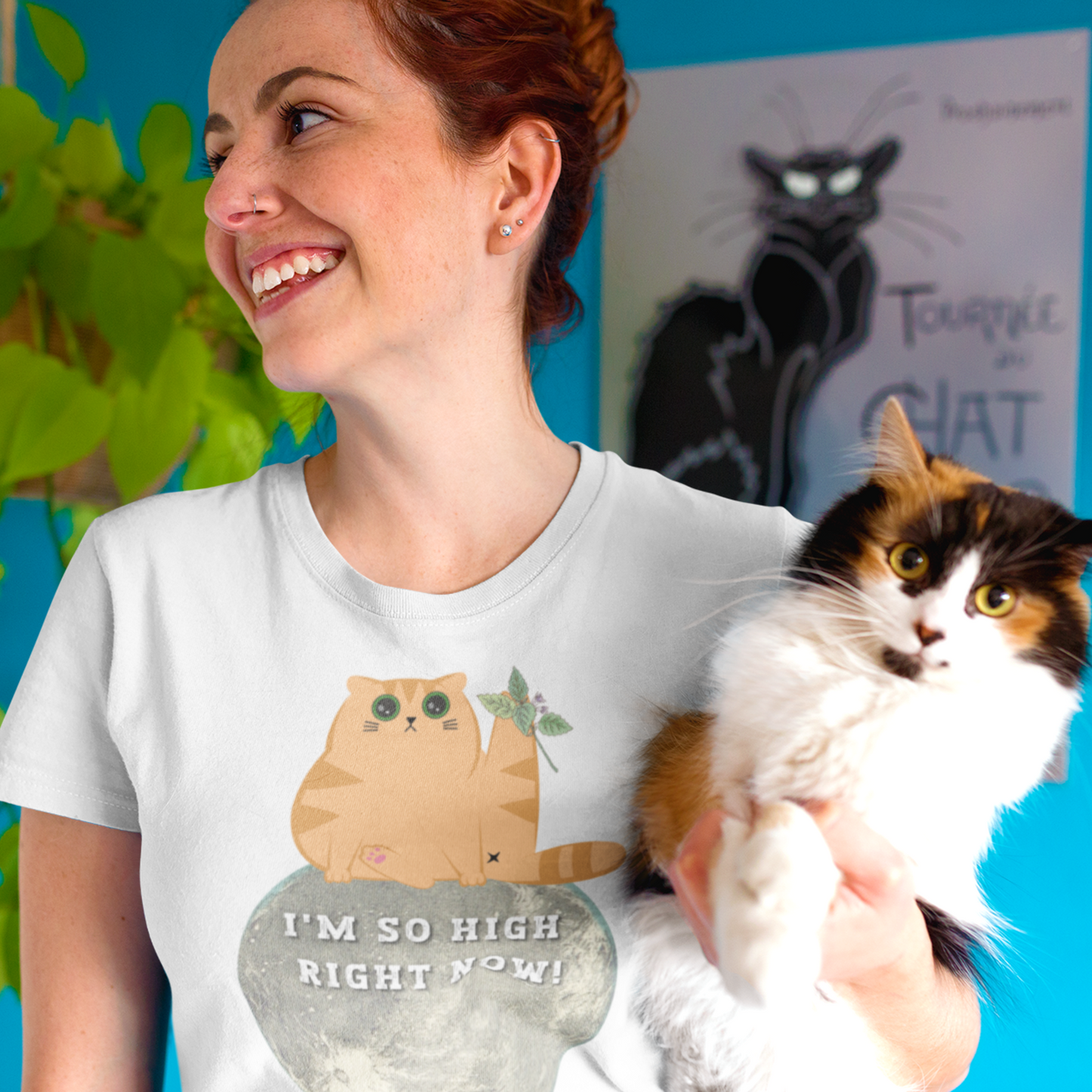 Young woman holding a cat wearing a white funny T-shirt featuring a ginger cat sat on the moon with wide eyes with catnip that reads 'I'm so high right now!'