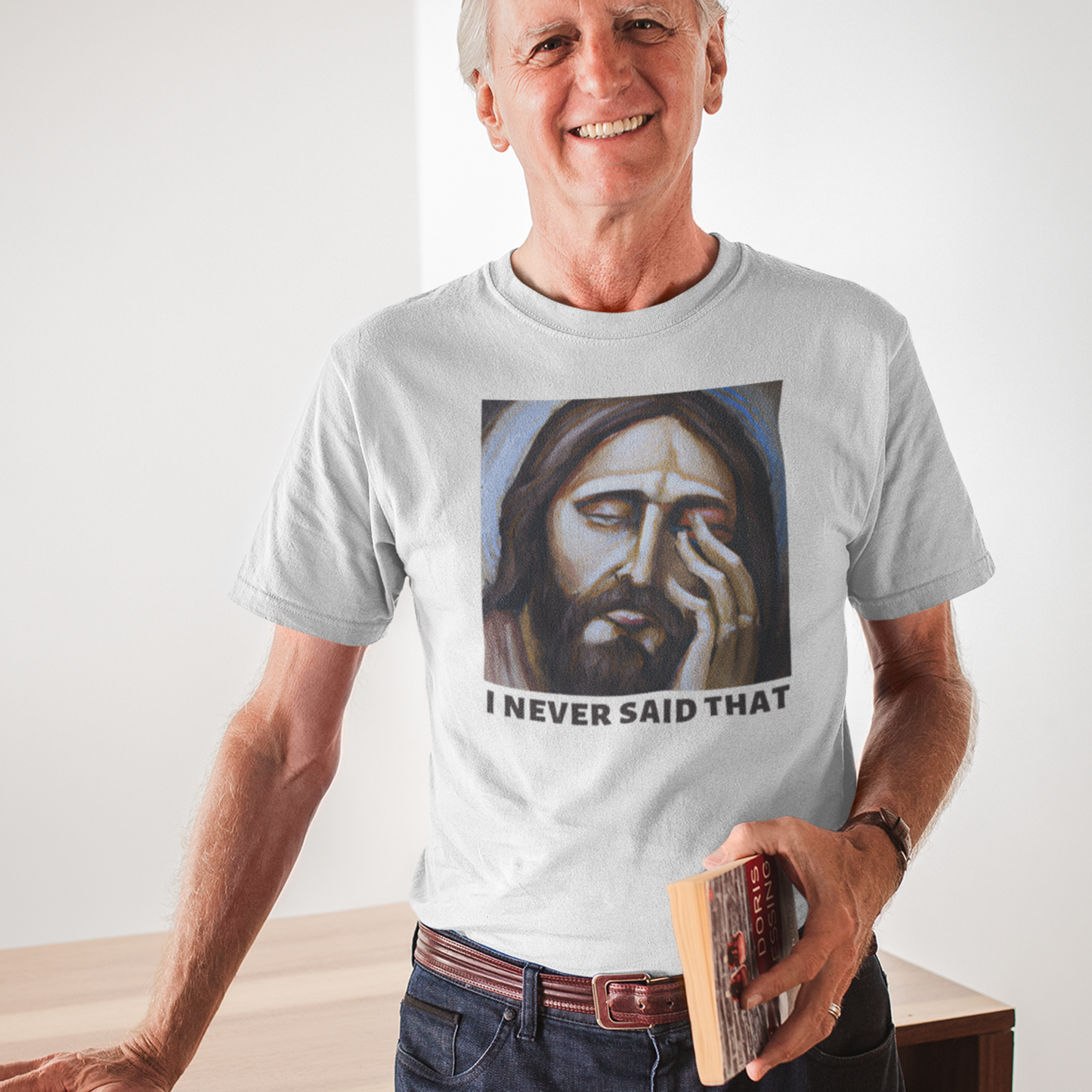 Distinguished old man holding a book wearing a white t-shirt with a graphic of Jesus with his hand over his face that reads I never said that