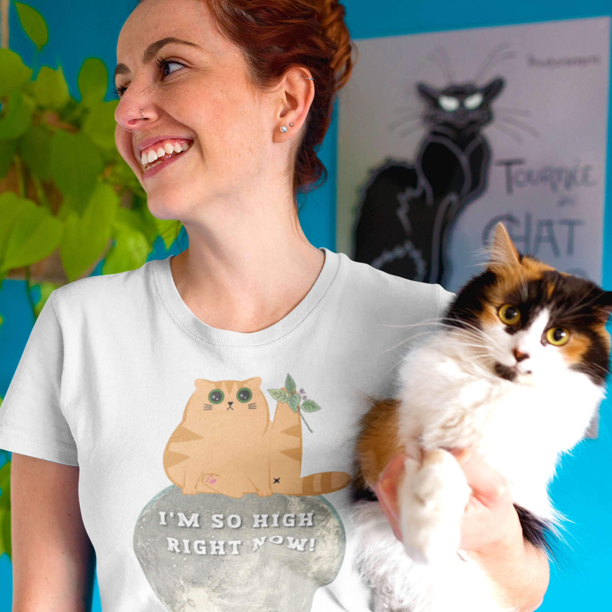 Young woman holding a cat wearing a white funny T-shirt featuring a ginger cat sat  on the moon with wide eyes with catnip that reads 'I'm so high right now!'