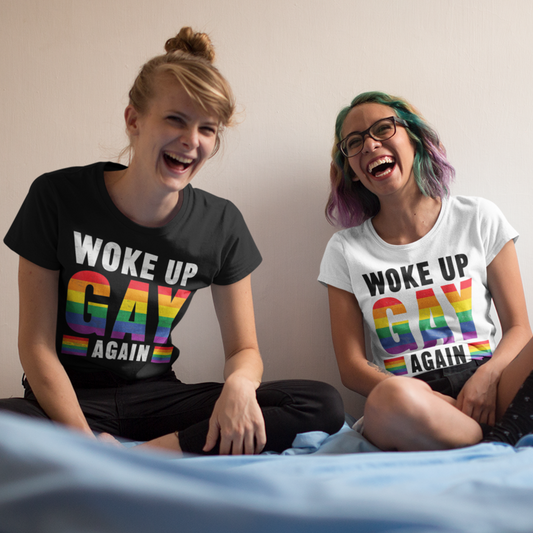 Two women laughing sitting on a bed wearing a t-shirt that reads Woke Up Gay Again in pride colours