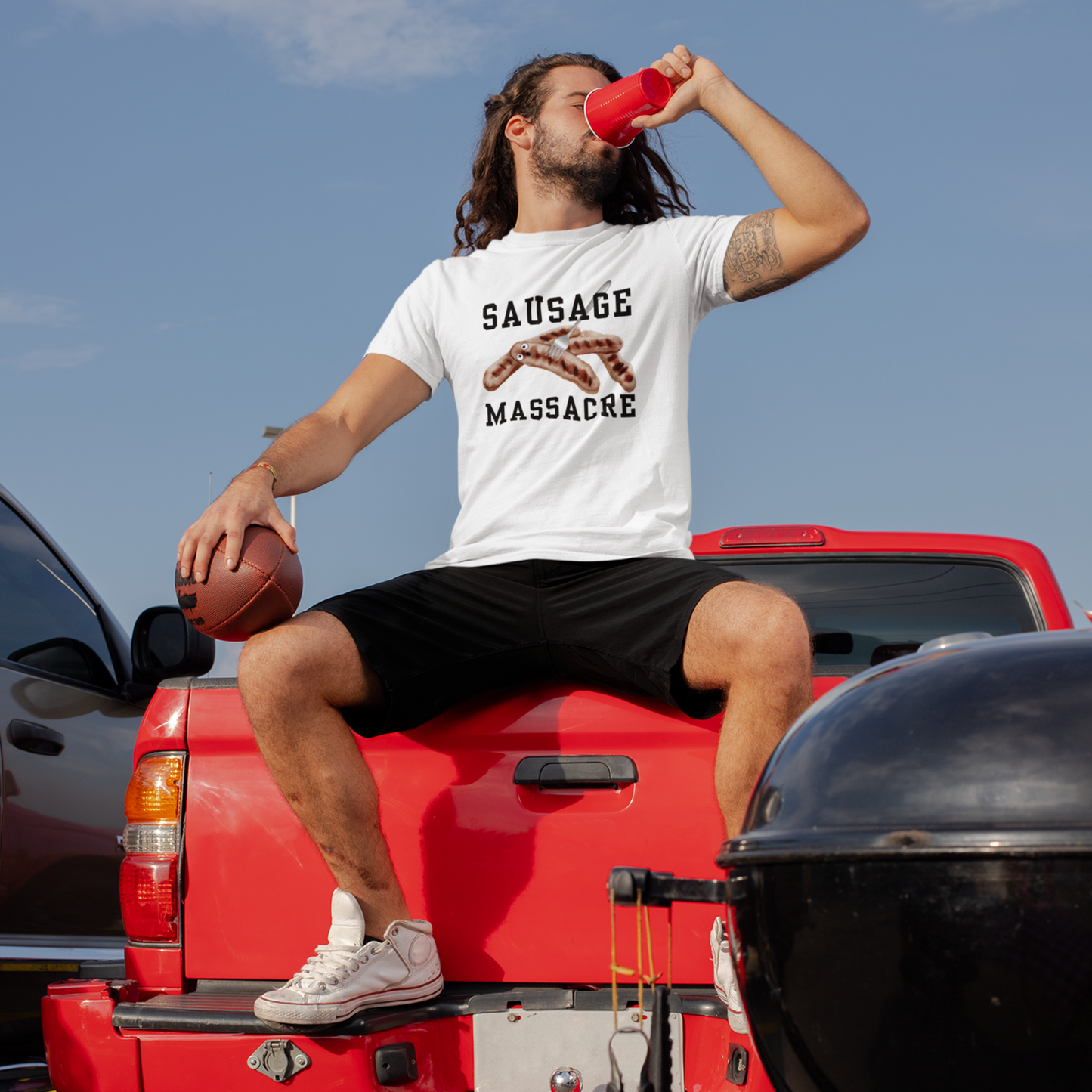 Young man sitting on a car drinking and wearing a white funny sausage massacre T-shirt with a fork through a shocked sausage.