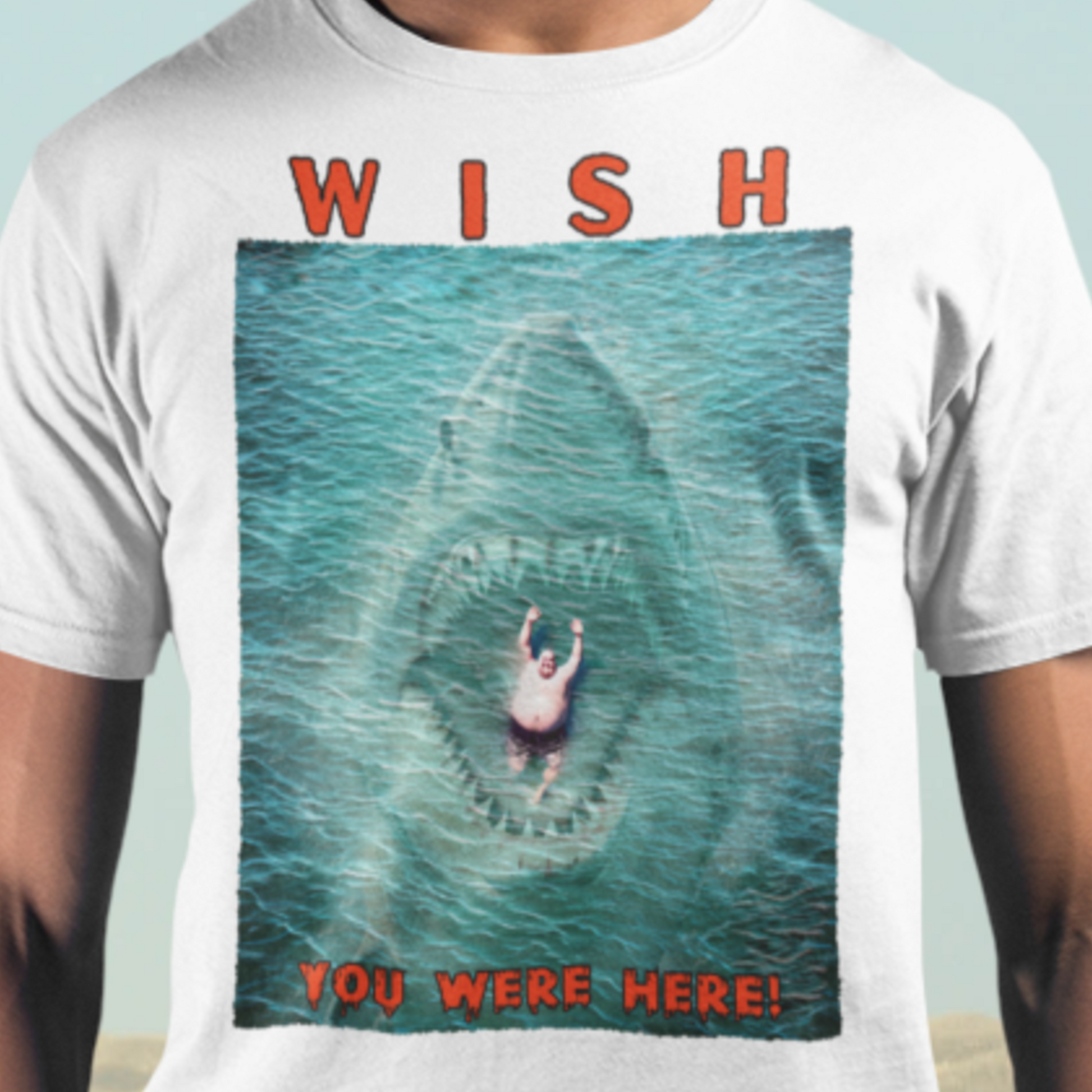 White, funny T-shirt with an image of a fat guy floating in the sea, with a large shark with its jaws open coming up underneath him, text reads, Wish you were here